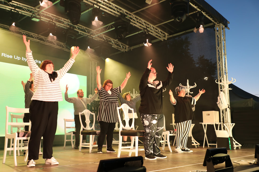 A group of nine actors on stage on a purpose built outdoor stage at Stockton International Riverside Festival. All are standing in front of white chairs, and are wearing a mix of black and white striped designed costume. They have both arms raised aloft in triumph. 
