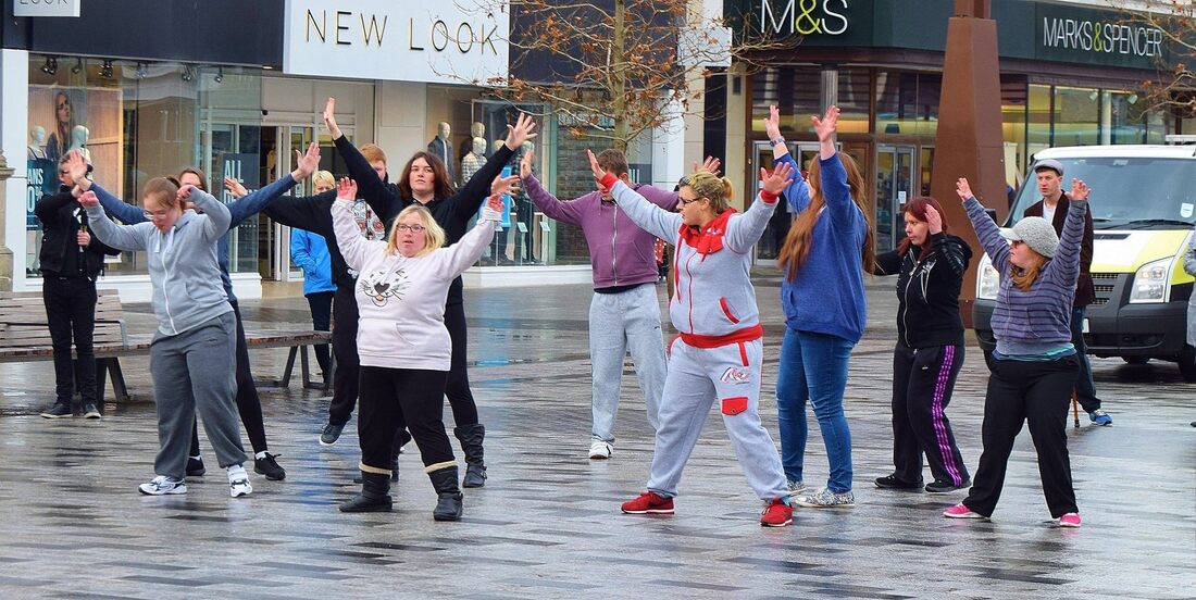 This photograph is full of energy as 11 young learning disabled people are dancing as part of a flash mob in the middle of Stockton High Street. All are wearing leisure and casual clothes and are standing with their arms in the air looking triumphant. High street shops are around them. 