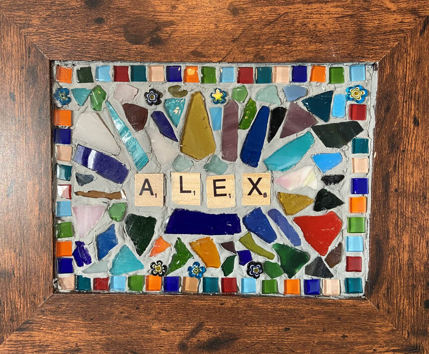 Mosaic by Alex Dechbamrung. The mosaic kits were created by Little Cog inspired by work seen on the internet. Within a brown wooden picture frame is a mosaic of each participant's name, created using scrabble style letter tiles, in this case Alex. The mosaic is created from a number of small square glass tiles in a range of colours and broken pieces of glass, again in a range of colours, arranged decoratively and grouted in position. Some strong blues, oranges and greens mixed in.