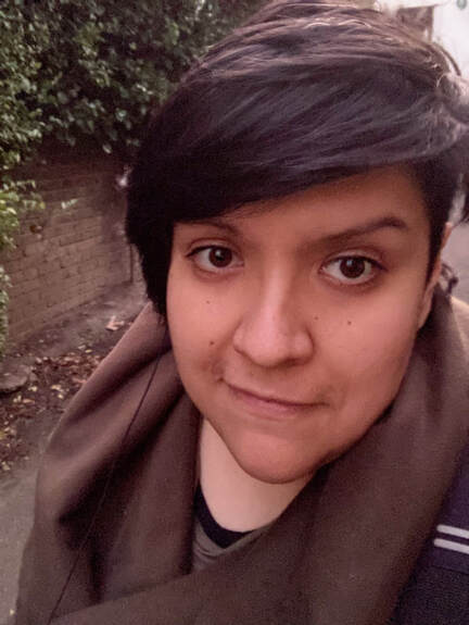 Cesia Leon-Alvarez is a young woman with short dark hair and is wearing a brown scarf and top. She is outdoors with a redbrish wall and green hedge behind her. 
