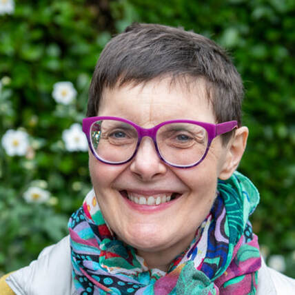 Caroline Mawer is an older white woman smiling broadly at the camera. She has very short dark hair, large glasses and is wearing a colourful scarf. She is outdoors and the background is leafy greenery. 