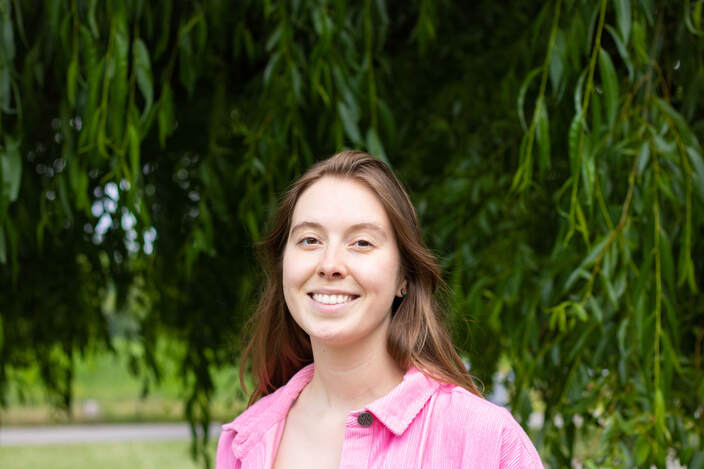 Elspeth Wilson is a young white woman with long brown hair smiling to camera, She is wearing a pink top and is outdoors standing under a large willow tree. 
