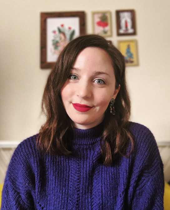 Lucy Goodwill is a young white woman with long dark hair, is smiling and wearing red liptsick as she smiles to camera. She is wearing a blue roll neck jumper and is seated in front of a wall of frame artworks. 