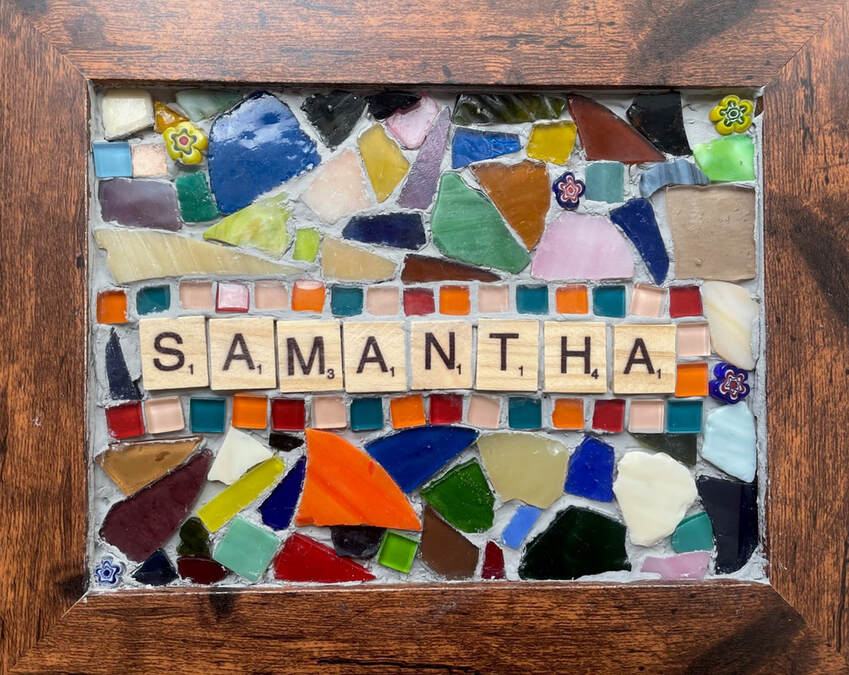 Mosaic by Samantha Blackburn. The mosaic kits were created by Little Cog inspired by work seen on the internet. Within a brown wooden picture frame is a mosaic of each participant's name, created using scrabble style letter tiles, in this case Samantha. There is a small square tile mosaic border to her name in orange, pink, teal and red. The other broken glass mosaic pieces are in various colours.