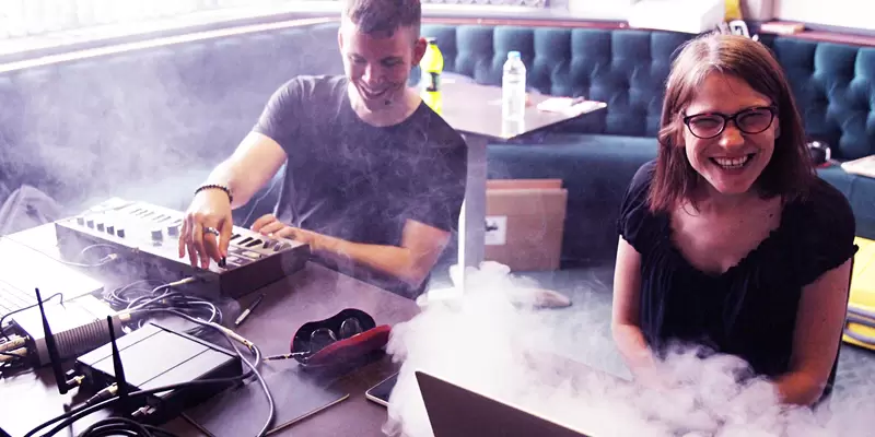 Gareth Cutter - a young white man with short brown hair, wearing a black tee shirt - and Gemma Nash - a young woman with a brown bob hairstyle and wearing dark rimmed glasses and a black top are seated at a sound desk surrounded by tech equipment with a special effect of smoke swirling. They are both laughing. 