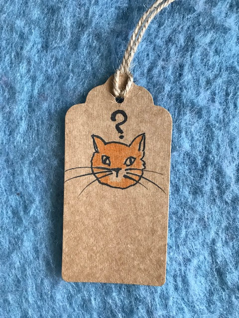 A brown parcel label on a blue wool background. It has a picture of a cat's face with a question mark above its head. Steph Fuller