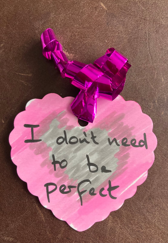 A pink heart label with the words I don't need to be perfect on a pink and grey background. Purple foil ribbon. 