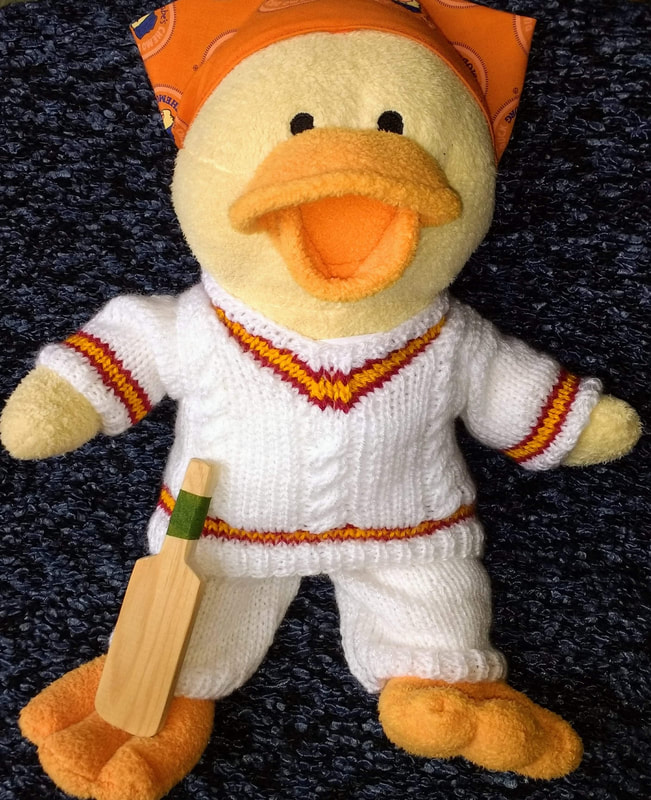 Image description - a soft toy cream coloured duck with yellow beak smiling. Lynne has knitted a cricket outfit of white trousers and a white V necked jumper with yellow and red stripe around the neckline, cuffs and waist. He has a little wooden cricket bat and is wearing a colourful head-covering.