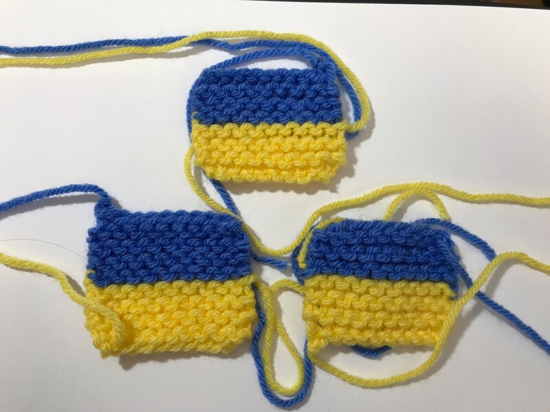 Three tiny knitted Ukrainian flags with blue and yellow yarn flowing away from them. Caroline Cardus. 