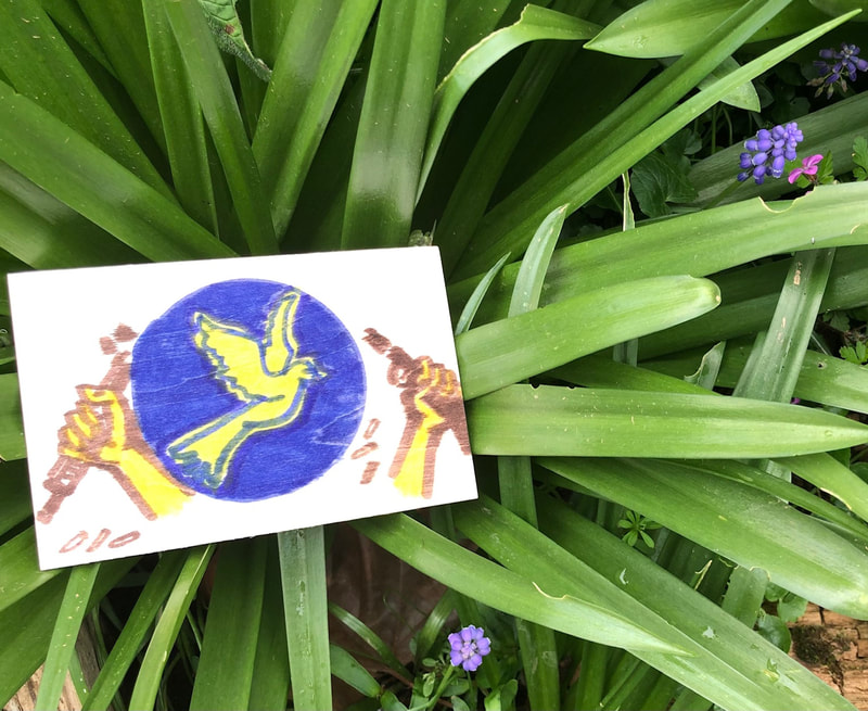 A painted panel with a yellow dove in a blue circle over which two hands are breaking a rifle, bullets falling to the floor. The panel is placed in greenery, long bluebell leaves and stems. JulieMc McNamara.