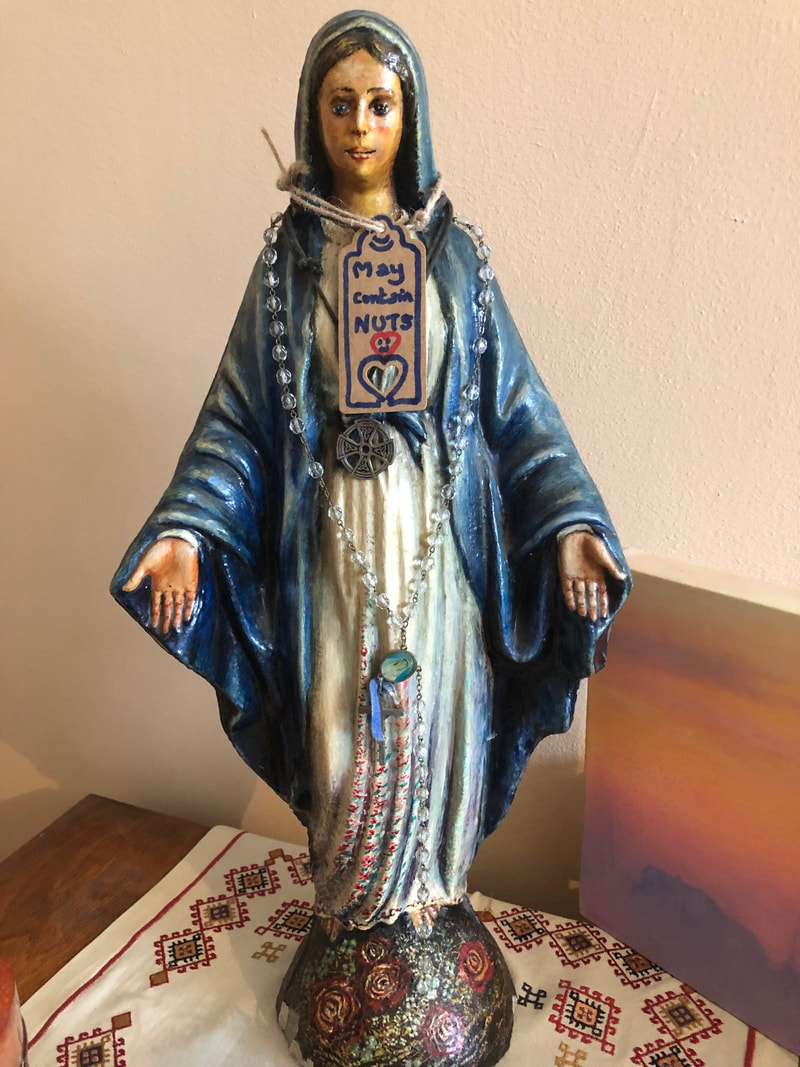 May Contain Nuts label on a string around a small statue of The Virgin Mary. She is wearing a blue cloak and has palms up turned and outstretched