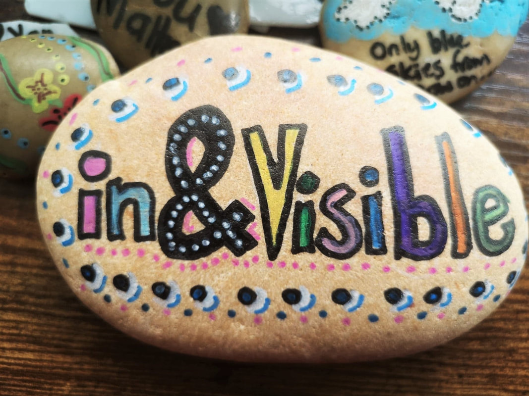 A hand decorated stone with the words in and visible in the centre. Colourful patterns of dots surround it. 