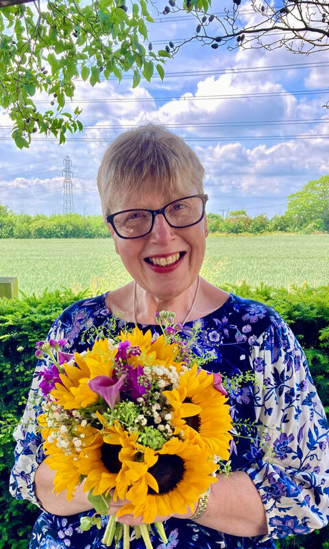 Lynne is a white woman with short blond hair and is wearing dark rimmed glasses and a blue floral top whilst holding a stunning posy of sunflowers. She is smiling warmly to camera with a blue sky and green fields background.