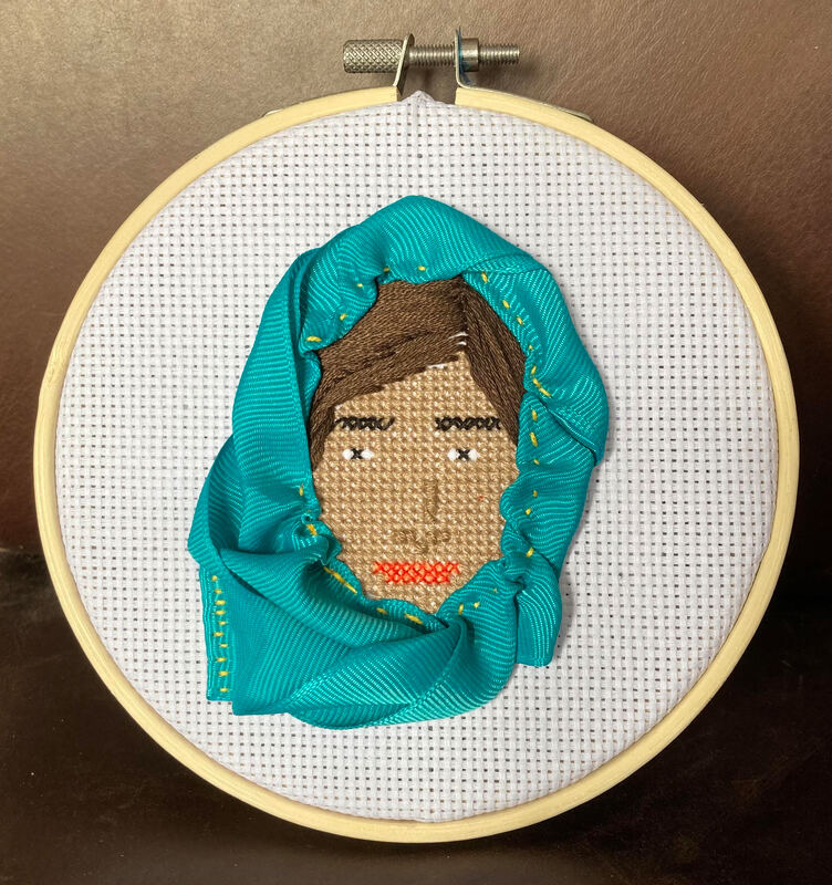 A cross stitch circular hoop containing a cross stitched portrait of Malala, a Pakistani activist for female education and the 2014 Nobel Peace Prize laureate. She survived being shot in the head during an assassination attempt when still a child because of her activism.  She is an Asian woman and in the cross stitch portrait id wearing a bright blue head covering. 