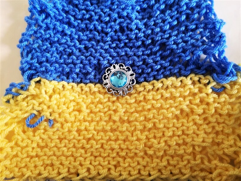 An irregular shaped, hand-knitted Ukrannian flag with a found jewel in the centre. Pale blue with a decorative silver metal surround. Pauline Heath.