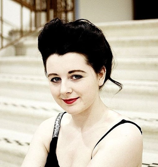 Porcelain Delaney is a young white woman with black hair styled up into a beehive. She is smiling to camera whilst standing at the bottom of some large steps outdoors. We can see the black and sequinned straps of a performance costume. 
