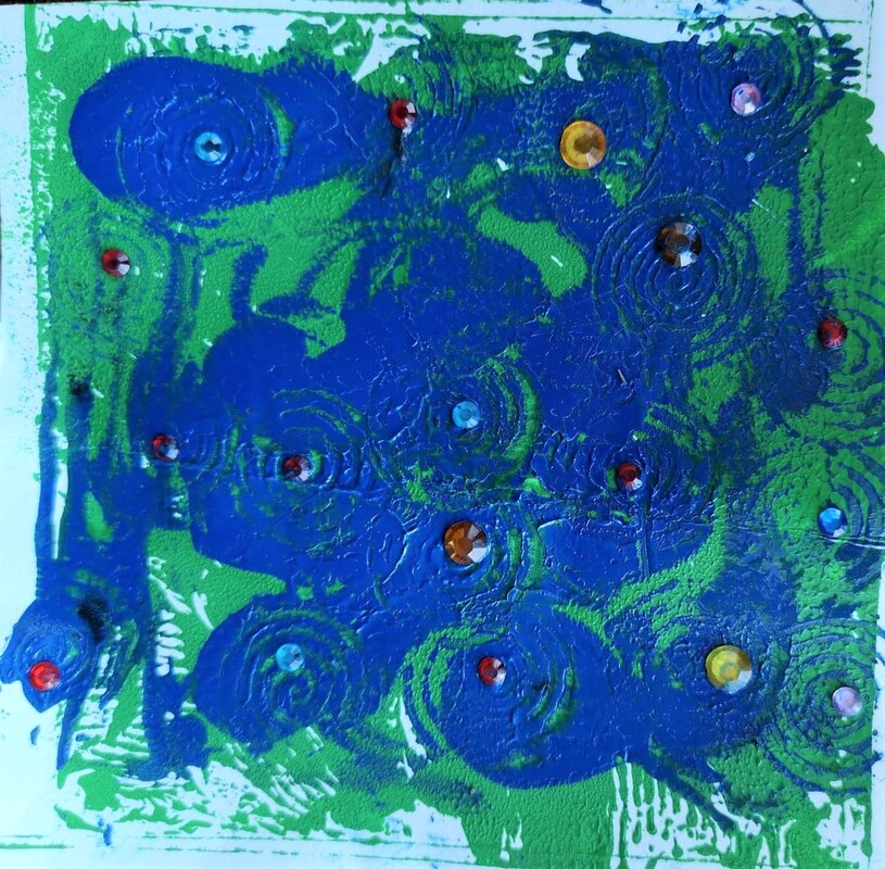 A pale blue background that has bright green stretched to the edges. A spiral wooden block has been used to add cobalt blue spirals on top of the bright green, some of the spirals are thick and almost solid and others are very thin and the spiral appears faded. Pauline has added different size jewels to the centre of the spirals. The jewels are different colours blue, red amber, yellow and pink.