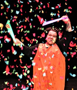 Image Description - Candice Keenan is a member of Full Circle. She is a young woman or mixed heritage with curly black hair, wearing a black top and a full length orange playsuit. She is dancing with glowsticks, and looks very happy as a confetti canon explodes above her, with confetti everywhere.