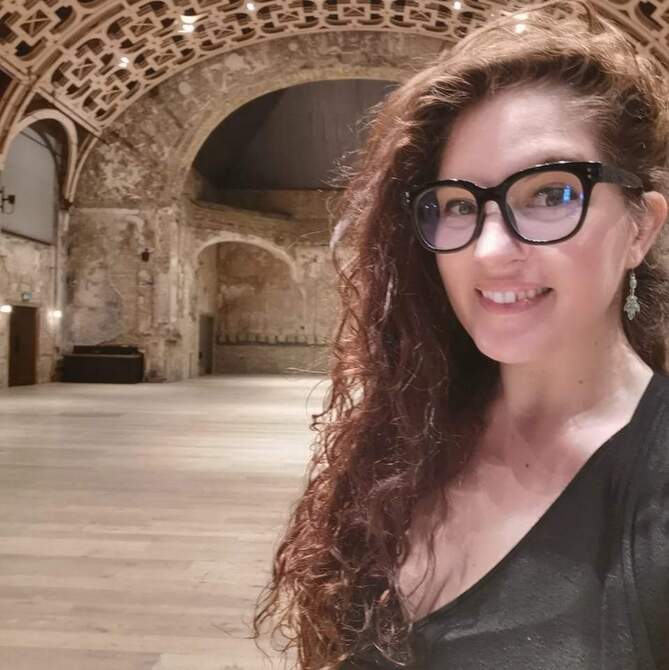 Deirdre is a white woman with long dark curled hair swept over to one side. She is wearing large dark rimmed glasses, and drop pendant earrings. She is wearing a black top, is smiling to camera and is standing in an old, large domed hall. 