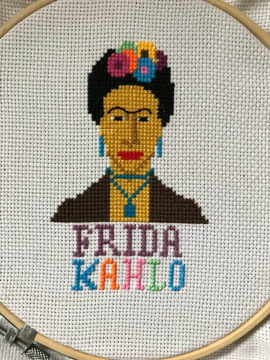 Cross stitch in an embroidery hoop - a portrait of Mexican artist Frida Kahlo. She is wearing a black top and head covering with several colourful flowers on it. She is also wearing blue earrings and pendant necklace.  The name Frida Kahlo is embroidered in different coloured yarns. 