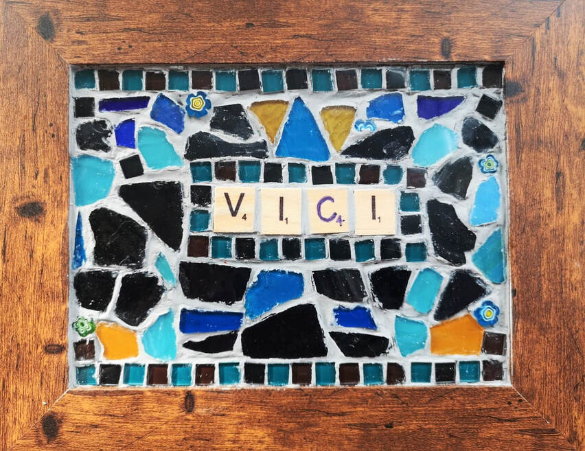 Mosaic by Vici Wreford-Sinnott.. The mosaic kits were created by Little Cog inspired by work seen on the internet. Within a brown wooden picture frame is a mosaic of each participant's name, created using scrabble style letter tiles, in this case Vici who you can tell has a history of cheating at Scrabble. She had to make her own letter C tile and awarded herself 4 points for it. There is a small square tile mosaic border to her name in black and teal. The other broken glass mosaic pieces are in various blues, black and four small gold pieces placed at the top and bottom of the pattern. 