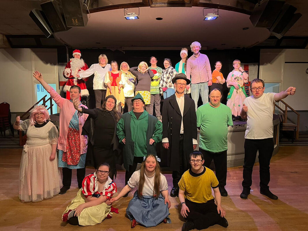 21 learning disabled actors in Christmas and pantomime costumes. Three people are seated on the floor at the front, a row of people is standing behind them, and then further behind, another row of people is standing on stage. They all look really pleased to be posing for their photo. 