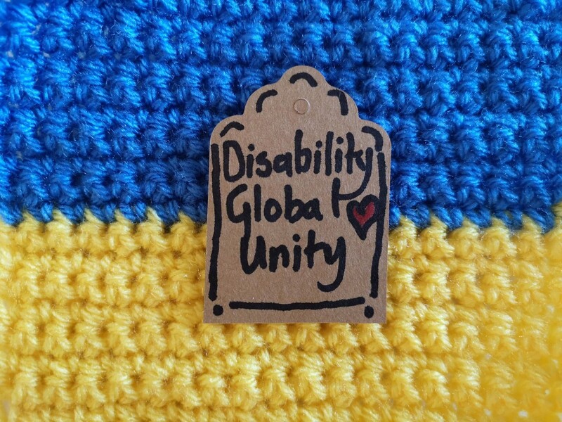 A crocheted Ukrainian flag with a label saying Disability Global Unity and a red heart. Vici Wreford-Sinnott
