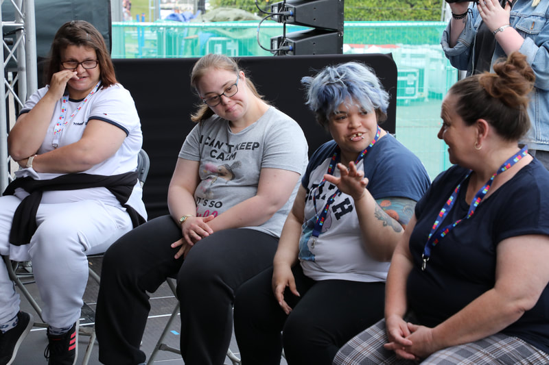 Four group members seated on stage at SIRF, chatting. Three brown haired white women and one woman of dual Caribbean and British heritage with blue hair. All wearing tee shirts.