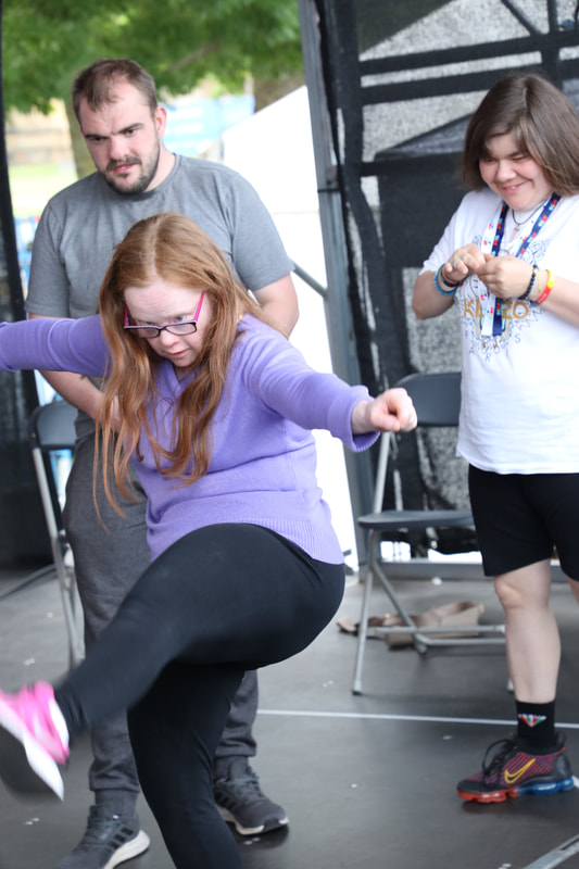 A young white woman with long red hair doing a high kick centre stage - others looking on and smiling wondering if they can do that.