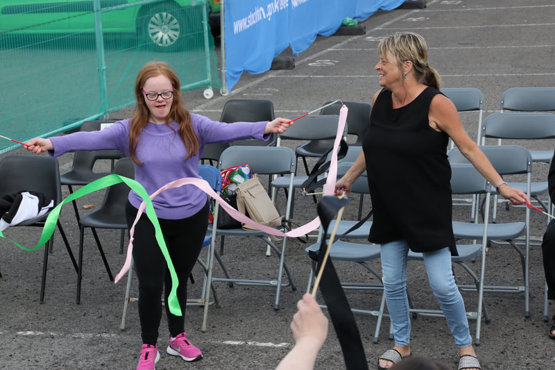 A young woman with long red hair and glasses doing ribbon work in the carpark workshop area, joined by a slightly older white woman. Both smiling. Colourful ribbons being twirled.