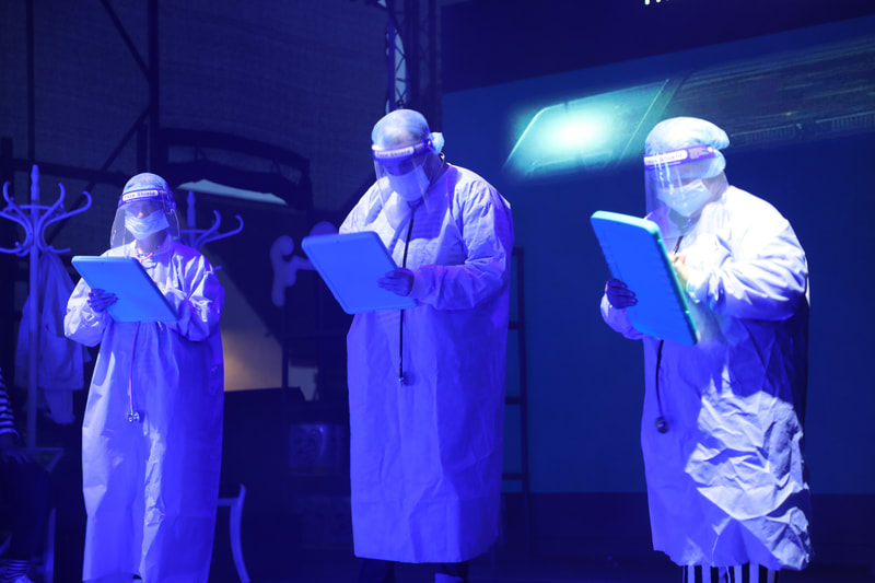 3 actors in a blue stage light, all wearing NHS scrubs gowns, blue hair nets, visors and masks, each has a stethoscope and a clip board. 
