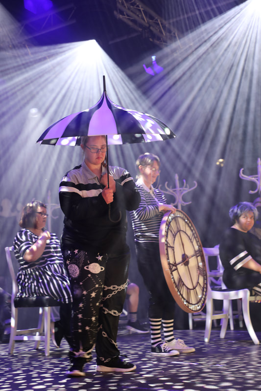 In the foreground a young female actor is standing under a black and whote umbrella, in the background an older female actor with a huge clock. Other actors seated. Dramatic lighting rays hitting the actors and stage. 