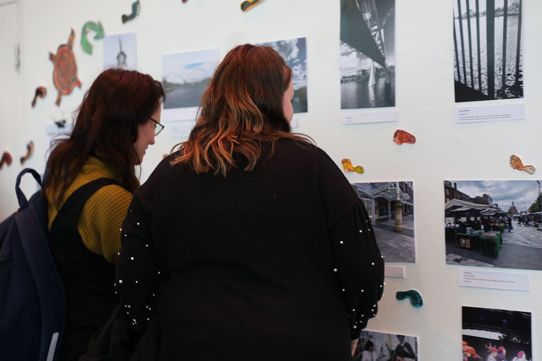 Two members of the public looking closely at a wall of photographs of local features of our stomping grounds. 