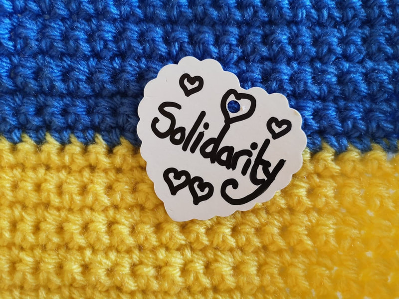 Crocheted Ukrainian flag with a white heart label on it reading 'solidarity' with five hand drawn hearts. Vici Wreford-Sinnott