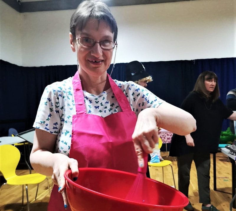 an older female actor wearing a red apron and using a red mixing bowl and whisk in a rehearsal in a dance studio. People in the background. 