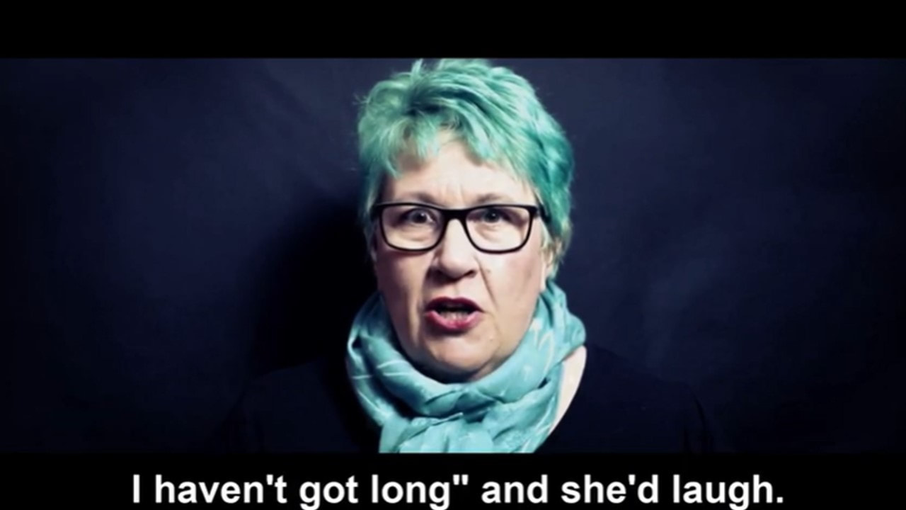 A screenshot of Vici Wreford-Sinnott performing to camera. She is a tall white woman in her fifties with teal hair, a teal scarf, dark top and dark rimmed glasses. Captions on the screen image read 'I haven't got long' and she'd laugh.