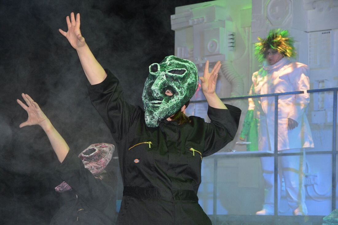 In the foreground is an actor actor wearing a black boiler suit and a large surreal handmade alien mask, standing boldly with arms in the air. Another actor in similar pose is behind them and on a  raised podium is a punk scientist character in customised white coat and green wig. 