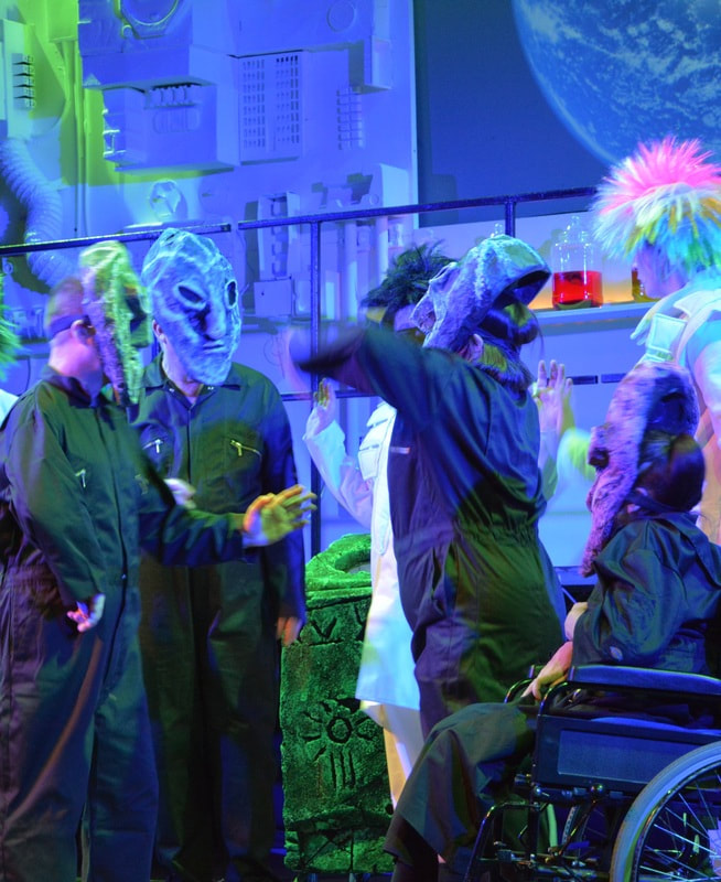 The second image also has colourful stage lighting and there are Four actors in the Lab, all in black boiler suits and large surreal handmade alien masks.