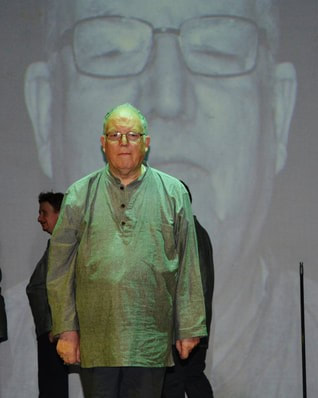 In the first image An older man is at the front of the stage, standing in an old fashioned institutional costume. A queue of people are behind his wearing the same outfit. His image is projected onto the screen behind as they were all having their photographs taken on arrival into the institution.