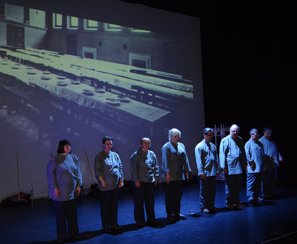 8 people are in a line at the front of the stage, all in an old-fashioned institution with a projected image of a dining hall with benches and tables. No one looks happy.