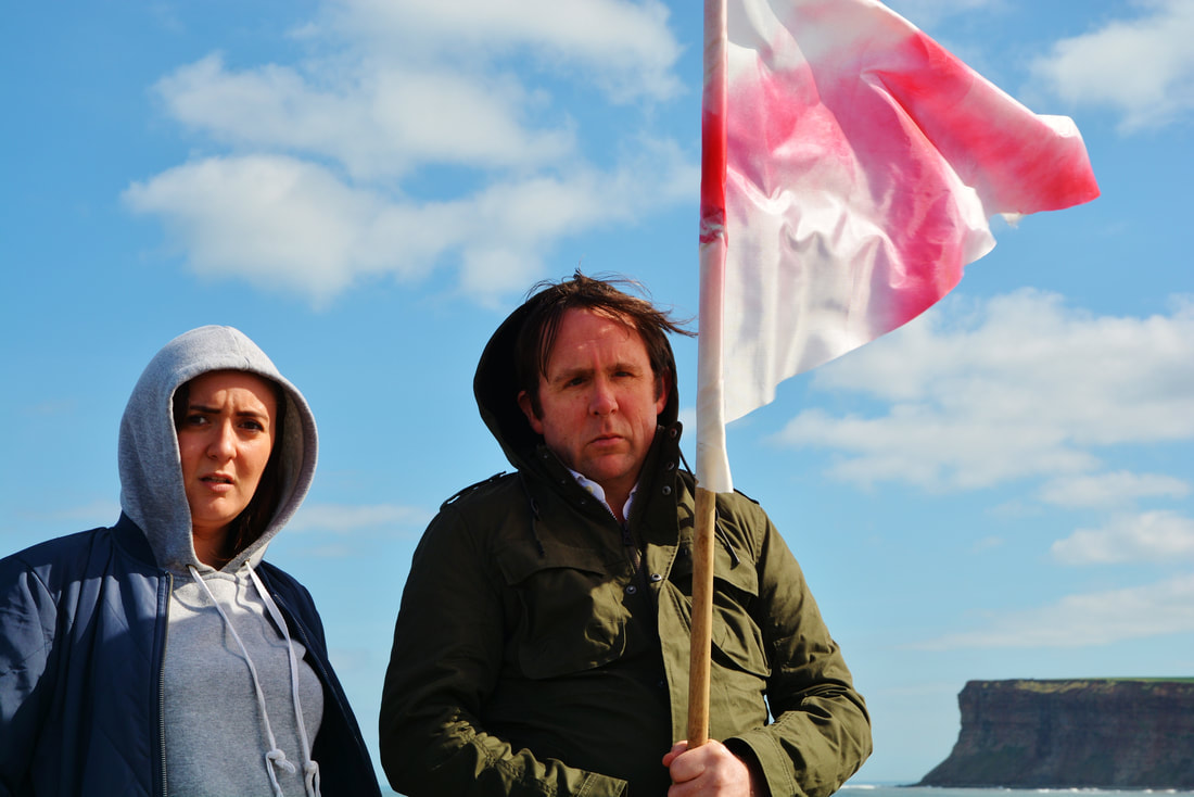 Two actors, Philippa Cole and Simon Startin stand in the foreground looking to the camera. They are outdoors with a blue sky as the backdrop and in the distance is a cliff from the North East coast. The edge of the country. Philippa plays Rat and Simon plays Murphy in Another England. Murphy is holding a hand made George's flag up.