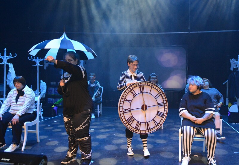 On stage oin atmospheric lighting actors in stylised black and white stripey costumes. One older female actor holding a huge clock and another younger female actor carrying a black and white umbrella