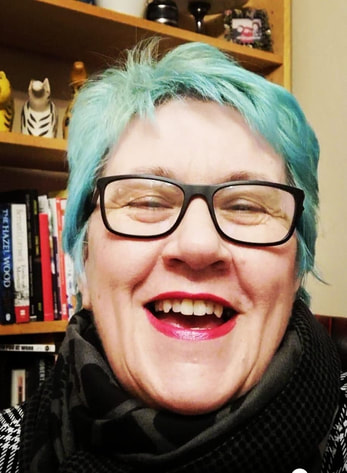Vici is a white woman in her fifties with short bright teal hair. She is wearing a dark rimmed glasses, a black scarf and a black and white checked top.  She is seated in front of a bookshelf and is laughing to camera.