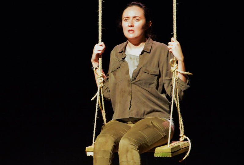 Actor Philippa Cole, a young white woman with ling dark hair in a pony tail is seated on a large stage swing. She is wearing Khaki green costume and looks concerned. 