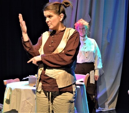 Image description - Actor Jennifer Lisky is in the foreground with long dark hair tied up in two small buns and wearing cream linen trousers and waistcoat and a brown grandad collar shirt, and actor Colly Metcalfe is in the background with long curly hair up in a bun and wearing a brown linen skirt with lines from the script sewn onto it in patches, and a cream linen shirt. Jennifer is playing Pip and Colly is playing Mona, a Deaf mother and daughter. The set is a large cream canvas drape at the back with lines of the script sewn onto it in patches and a dining table and two chairs in the middle of the stage. Jennifer is performing in BSL.