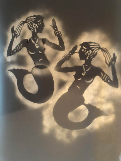 Two images of mermaids, white spray over charcoal. Handcut stencils with long hair, bangles, a pendant, earrings, and naked with bre3ats, nipples and large buttocks, and a mermaid's tail. 