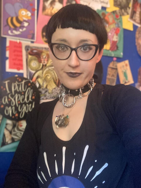 Jessica Secmezsoy-Urquhart is a young non-binary person with short black hair with a high fringe cut. They are wearing a black top and large dark rimmed glasses. They are also wearing a black leather choker with silver loops and an ornate silver necklace and pendant. Jessica is seated in front of a colourfull wall covered in postcards, art, and posters.