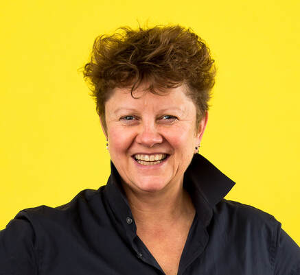 JulieMc McNamara is an older white woman beaming at the camera, ready for mischief. She is wearing a navy blue shirt and is against a bright yellow background. She has short brown hair. 