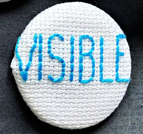 An embroidered badge by Lynne Mcfarlane. White embroidery fabric stretched over a badge with the word VISIBLE in blue yarn.