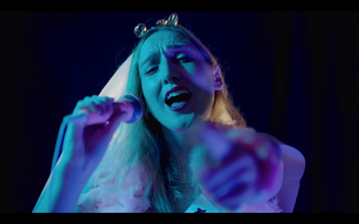 Nicola Chegwin in Hen Night for BBC i-Player. She is lit in blue lighting and is singing into a karaoke microphone. She is dressed in Hen Night garb and is reaching out to the camera smiling. 